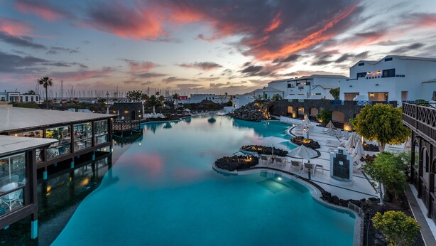 A resort pool with a sunset view, lounge areas, and a bridge connecting different sections. - Hotel LIVVO Volcán Lanzarote (Yaiza)