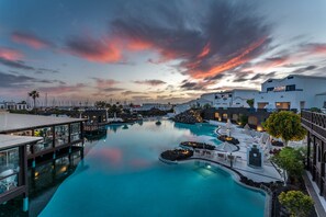 Een resort zwembad met uitzicht op de zonsondergang, met loungegedeeltes en een brug die verschillende zones verbindt.