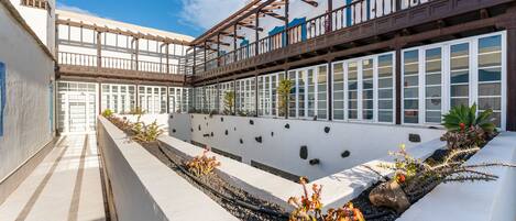A courtyard with a white wall, a wooden pergola, and a row of windows.