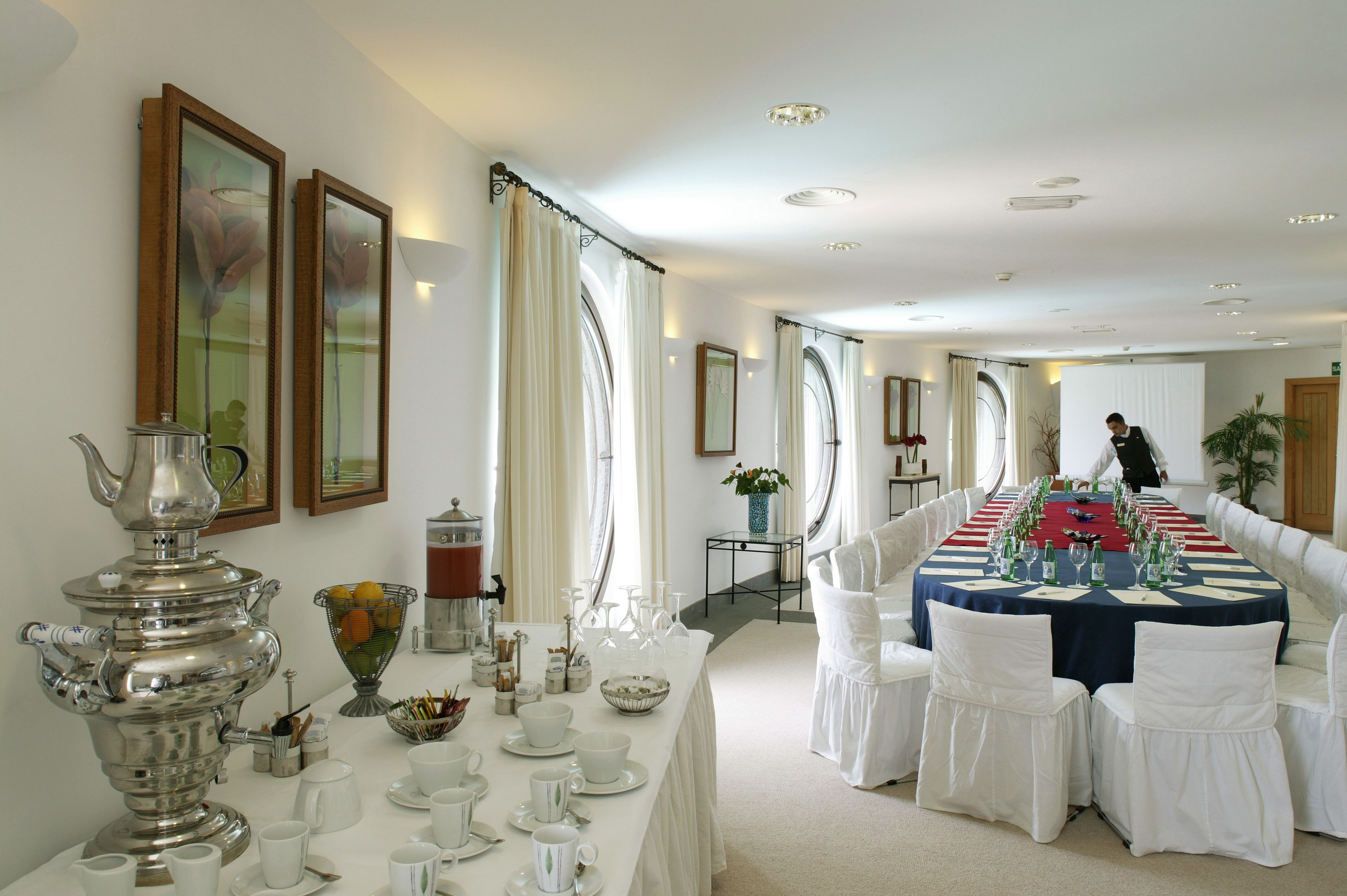 A formal dining room with a long table set for a meal, white chairs, and a silver samovar on the left.