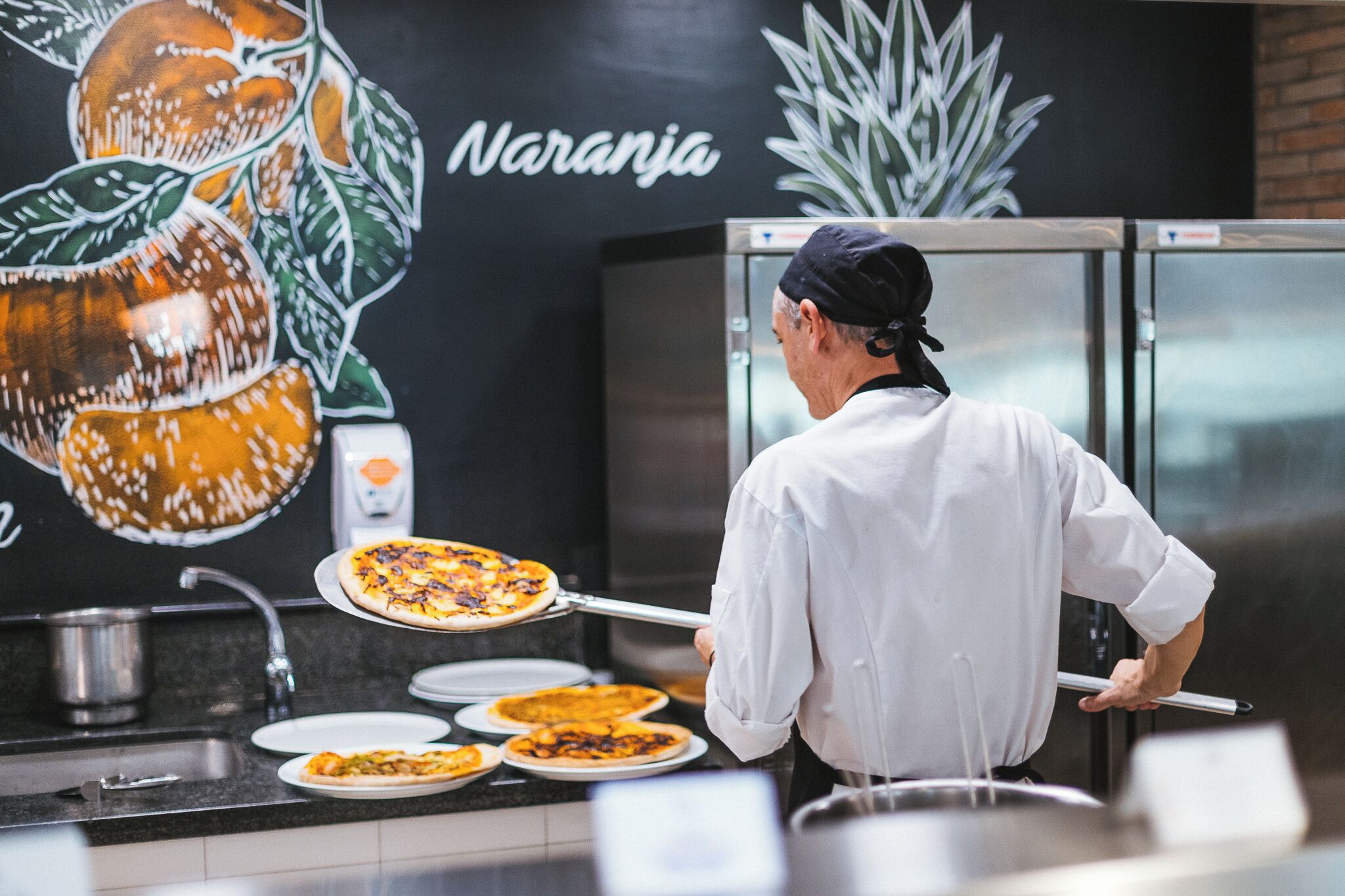 A chef in a white uniform and black cap preparing pizzas in a kitchen with a mural of fruits and the word "Naranja" on the wall.