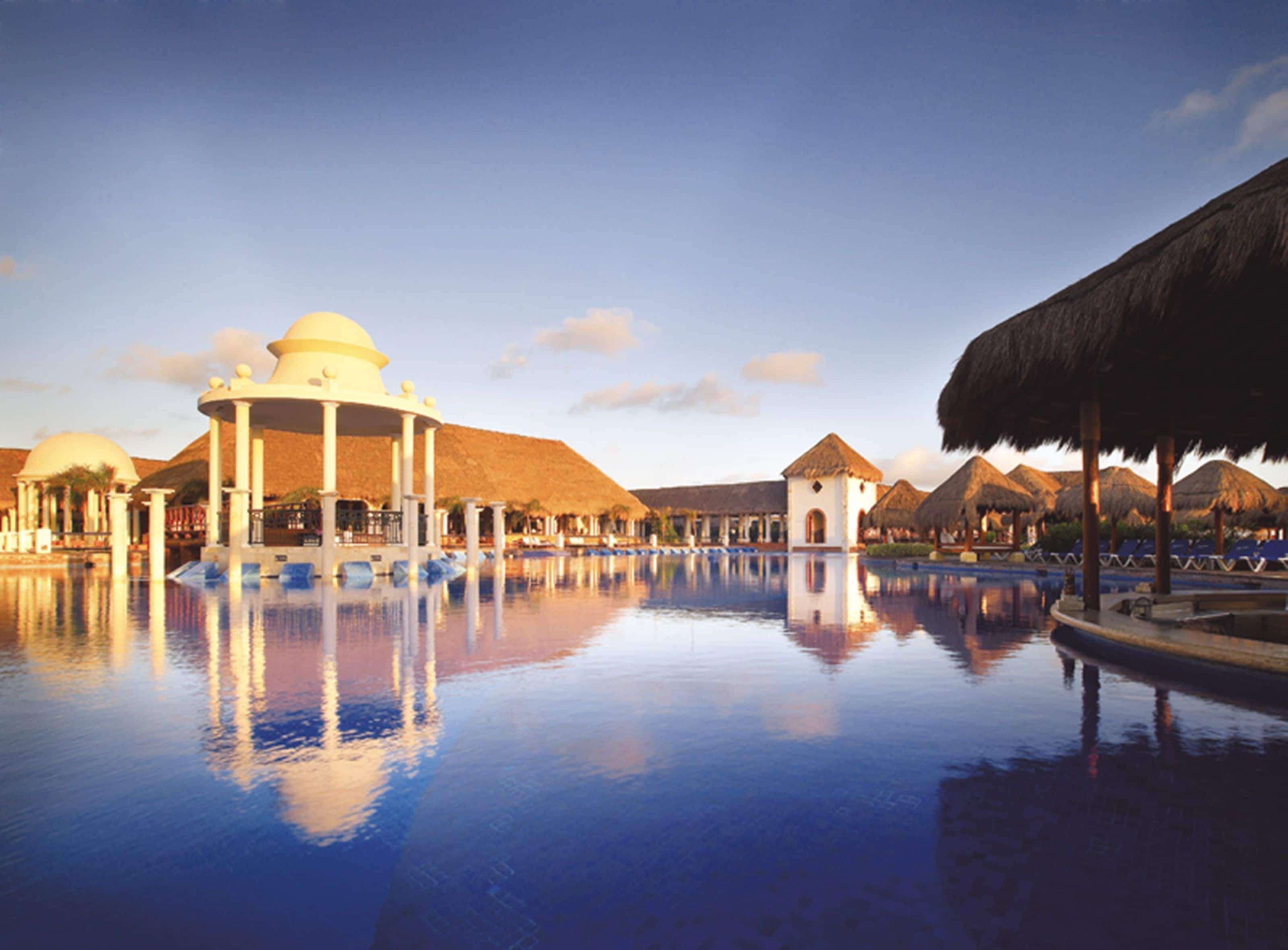 Piscina de resort con reflejo claro, rodeada de construcciones con techo de paja y un edificio central con cúpula.