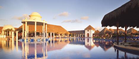 Piscina de resort con reflejo claro, rodeada de construcciones con techo de paja y un edificio central con cúpula.