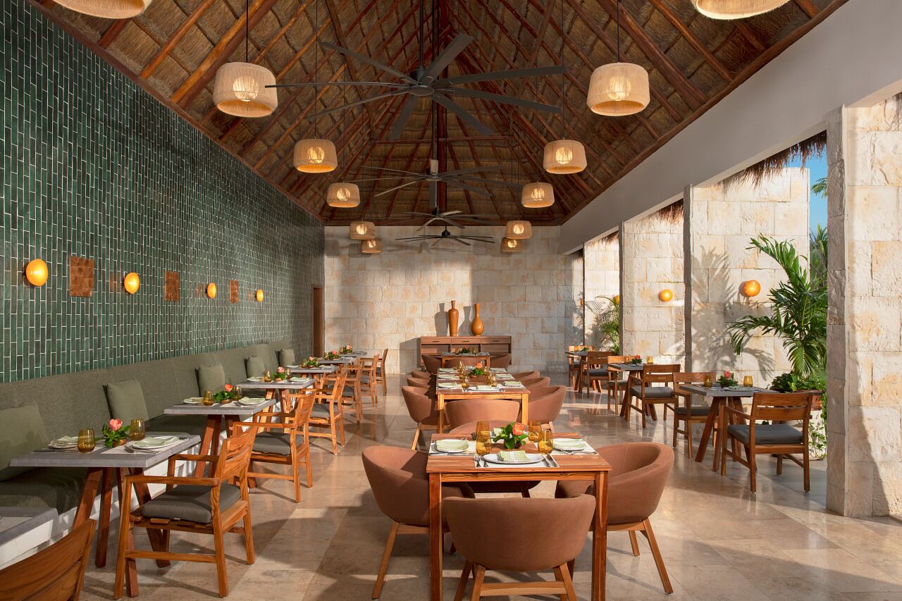A restaurant with wooden tables and chairs, a high ceiling with a fan, and a green tiled wall.