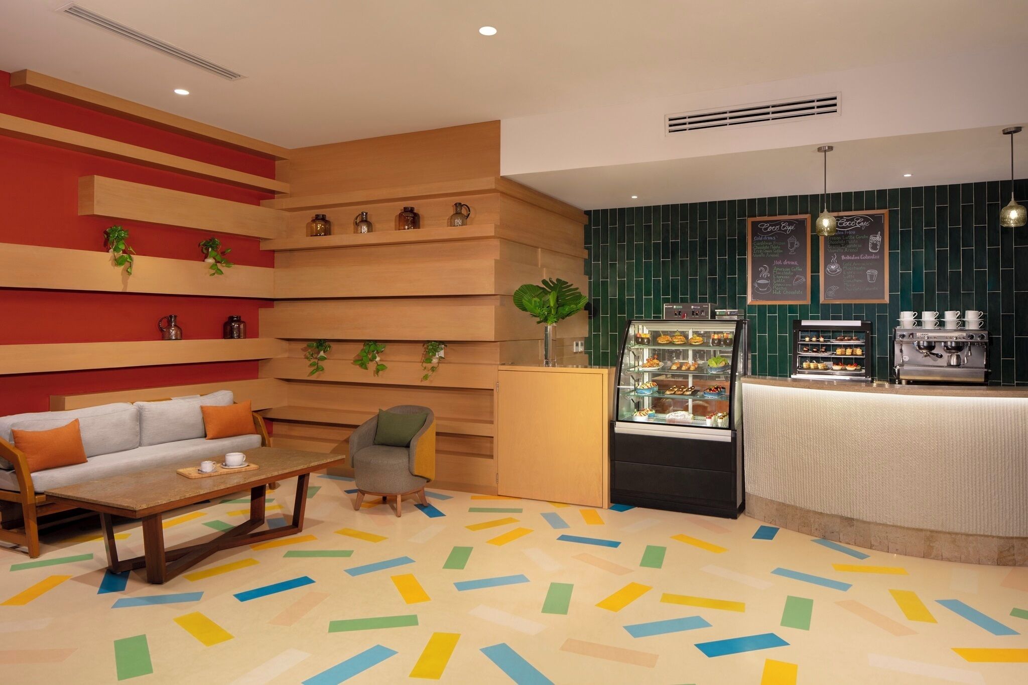 Un café moderne avec un comptoir d’accueil en bois, un bar avec une machine à café et une vitrine remplie de pâtisseries. On y trouve un canapé et une petite table avec des tasses et des soucoupes.