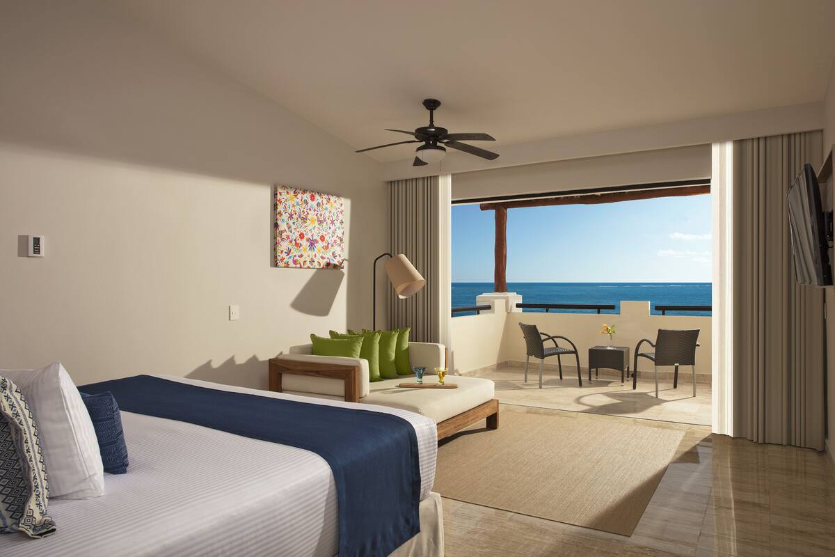 Een hotelkamer met een bed, een plafondventilator, een balkon met uitzicht op zee, en een tafel met stoelen.