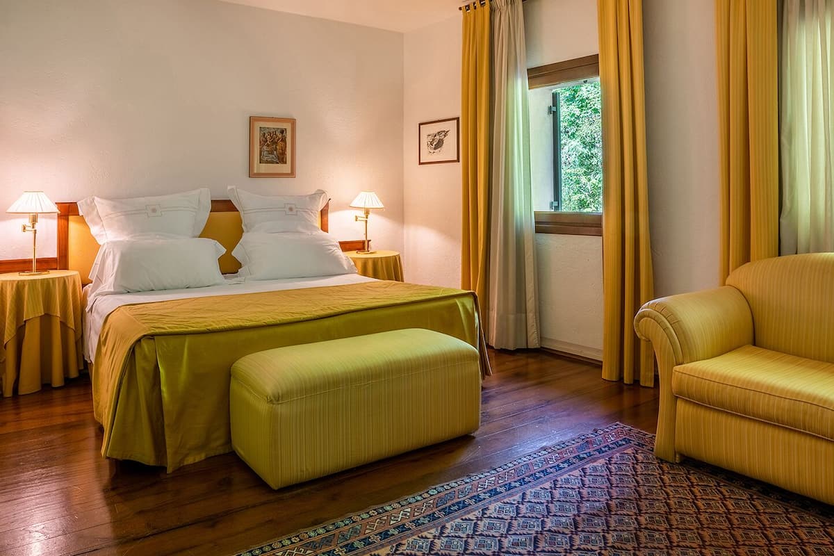 Una habitación de hotel con una cama, dos lámparas de noche, un ventanal con cortinas, un sofá y una alfombra estampada.