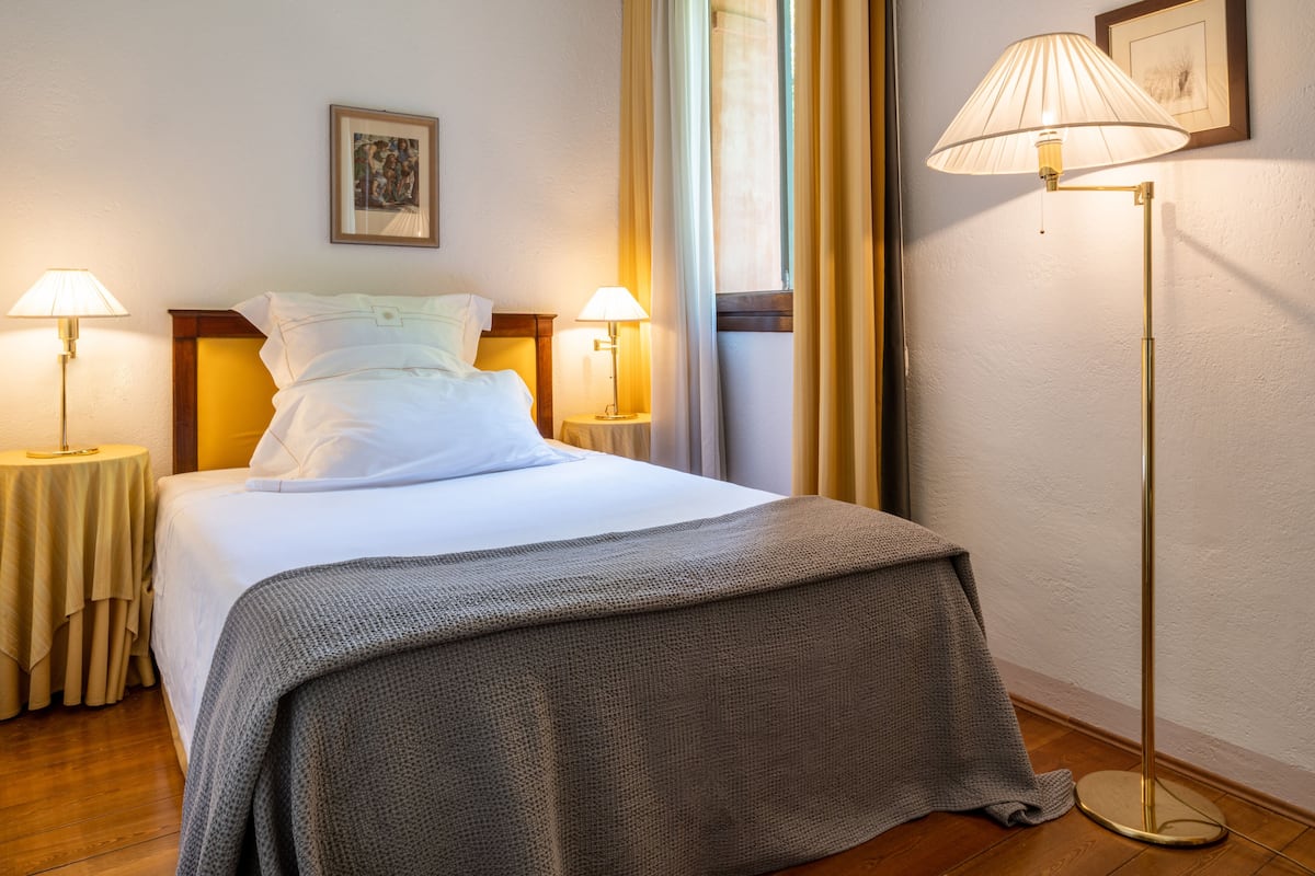 Una cama bien hecha con una manta gris, dos lámparas de noche, un cuadro enmarcado en la pared y una lámpara de pie.