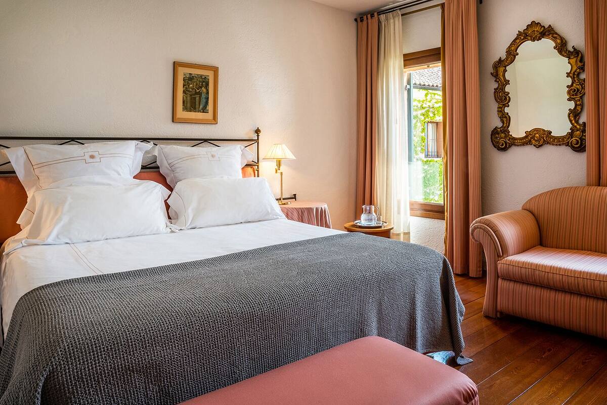Una cama bien hecha con sábanas blancas, un edredón estampado en gris y negro, una mesita de noche de madera con una lámpara, un cuadro en la pared y una ventana con cortinas.