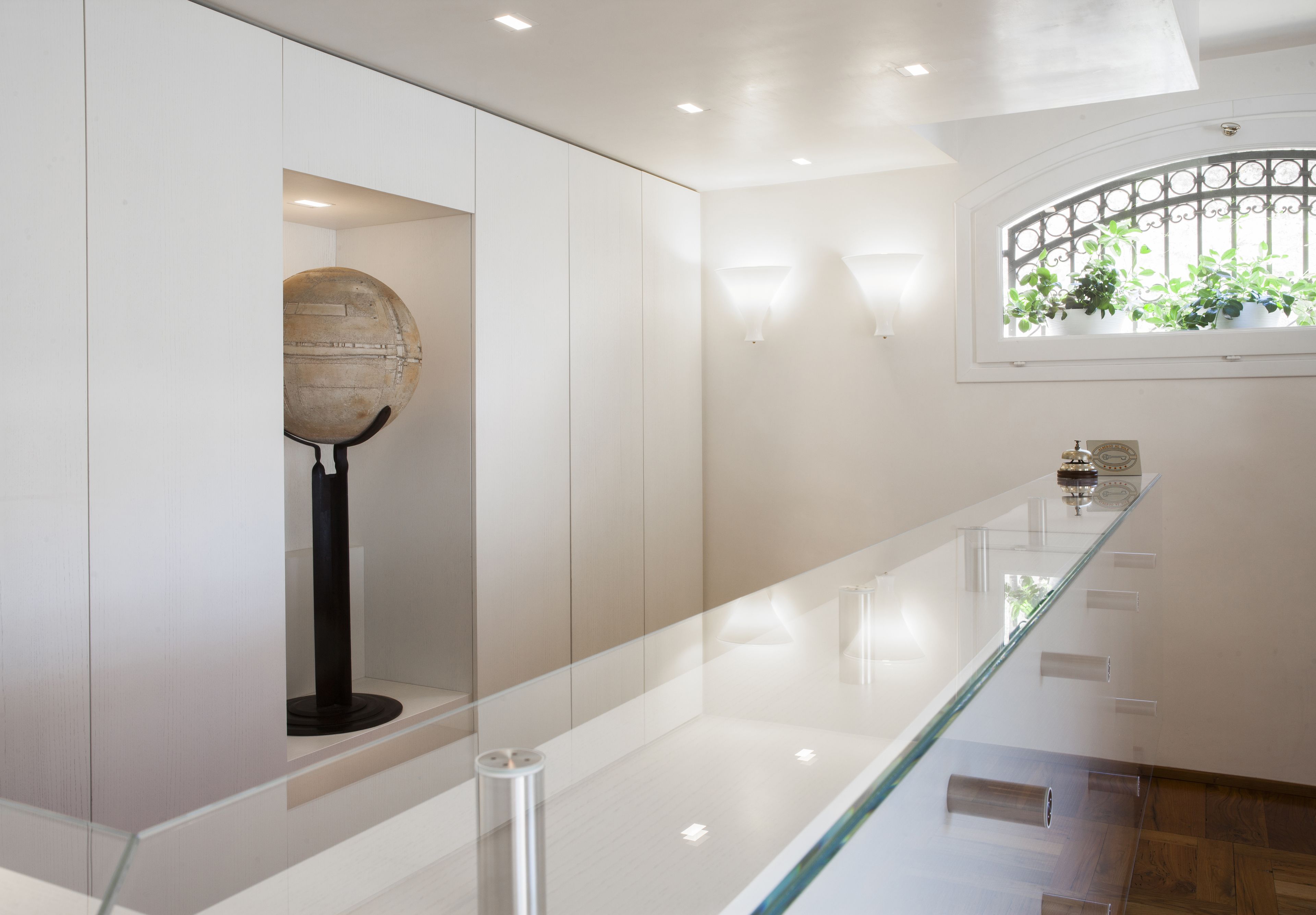 A modern interior with a glass railing, a spherical sculpture on a pedestal, and a window with plants.