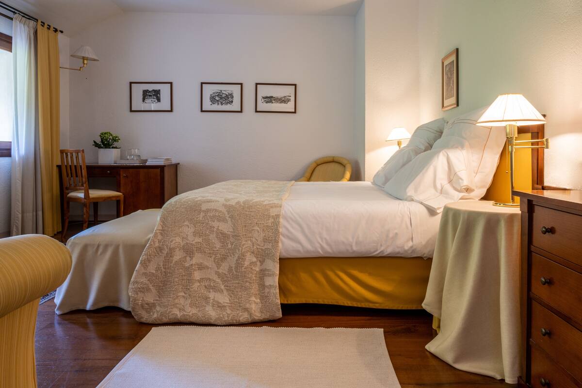 Un dormitorio ordenado con cama, mesitas de noche, una silla y un escritorio. Hay dos cuadros en la pared.