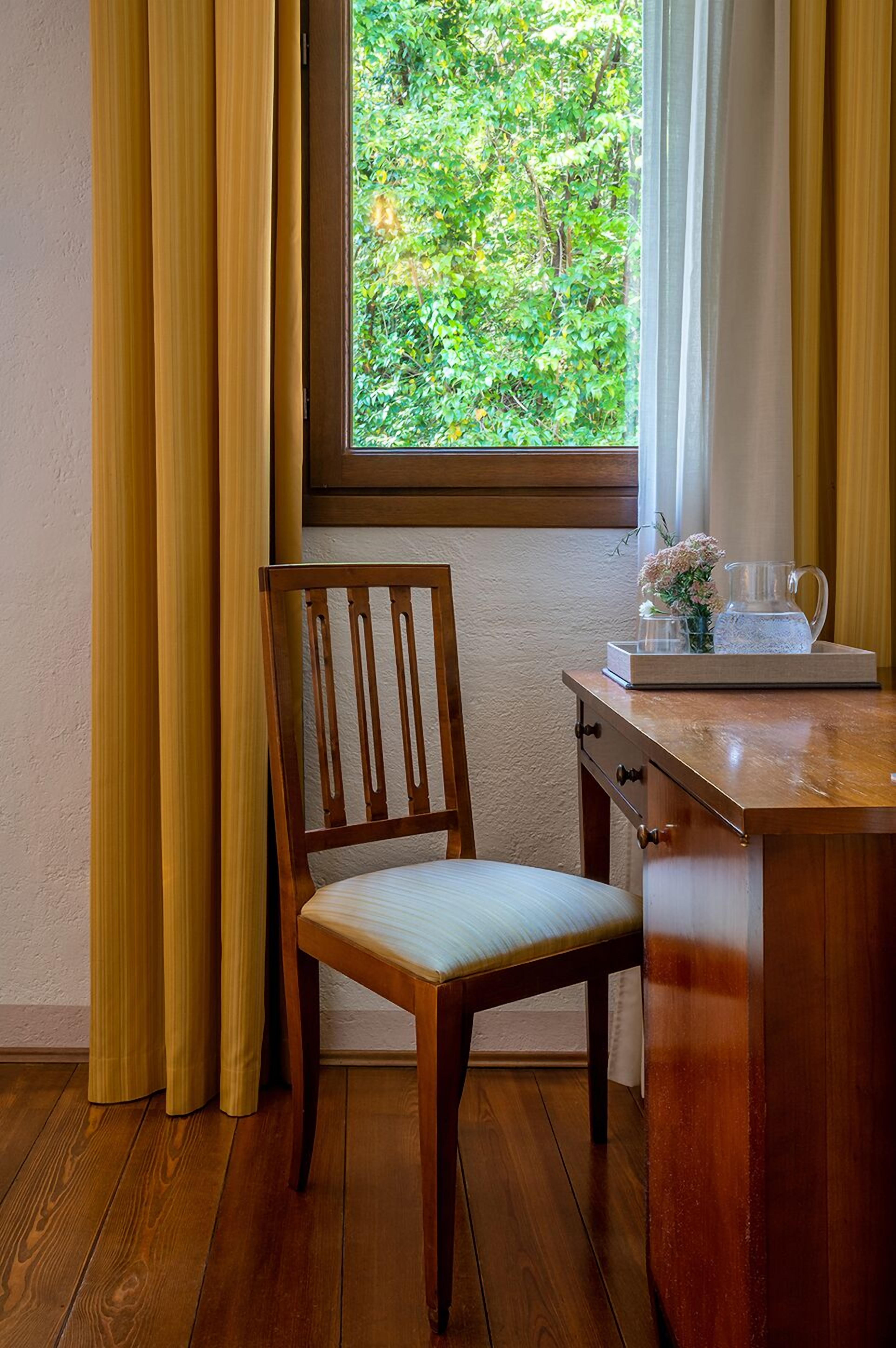 A wooden chair with a cushioned seat and backrest, positioned next to a wooden desk with drawers. A window with a view of greenery is visible in the background.