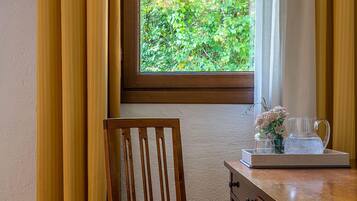 Una silla de madera con asiento y respaldo acolchados, ubicada junto a un escritorio de madera con cajones. Se ve una ventana con vistas a la vegetación al fondo.