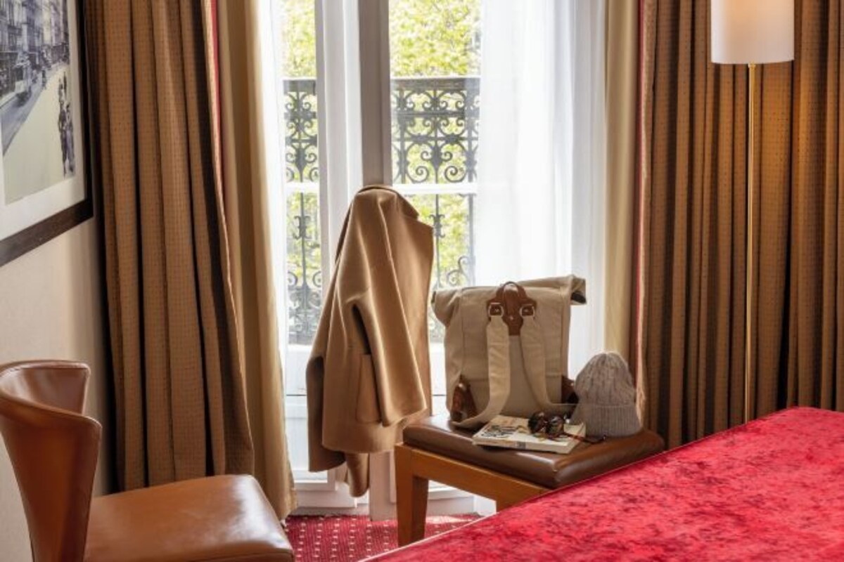A hotel room with a bed, a chair, a table with a handbag and a hat, and a window with curtains.