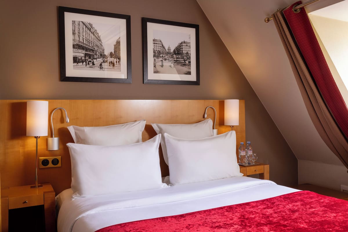 A hotel room with a large bed, two framed pictures on the wall, a wooden headboard, bedside lamps, and a red throw blanket.