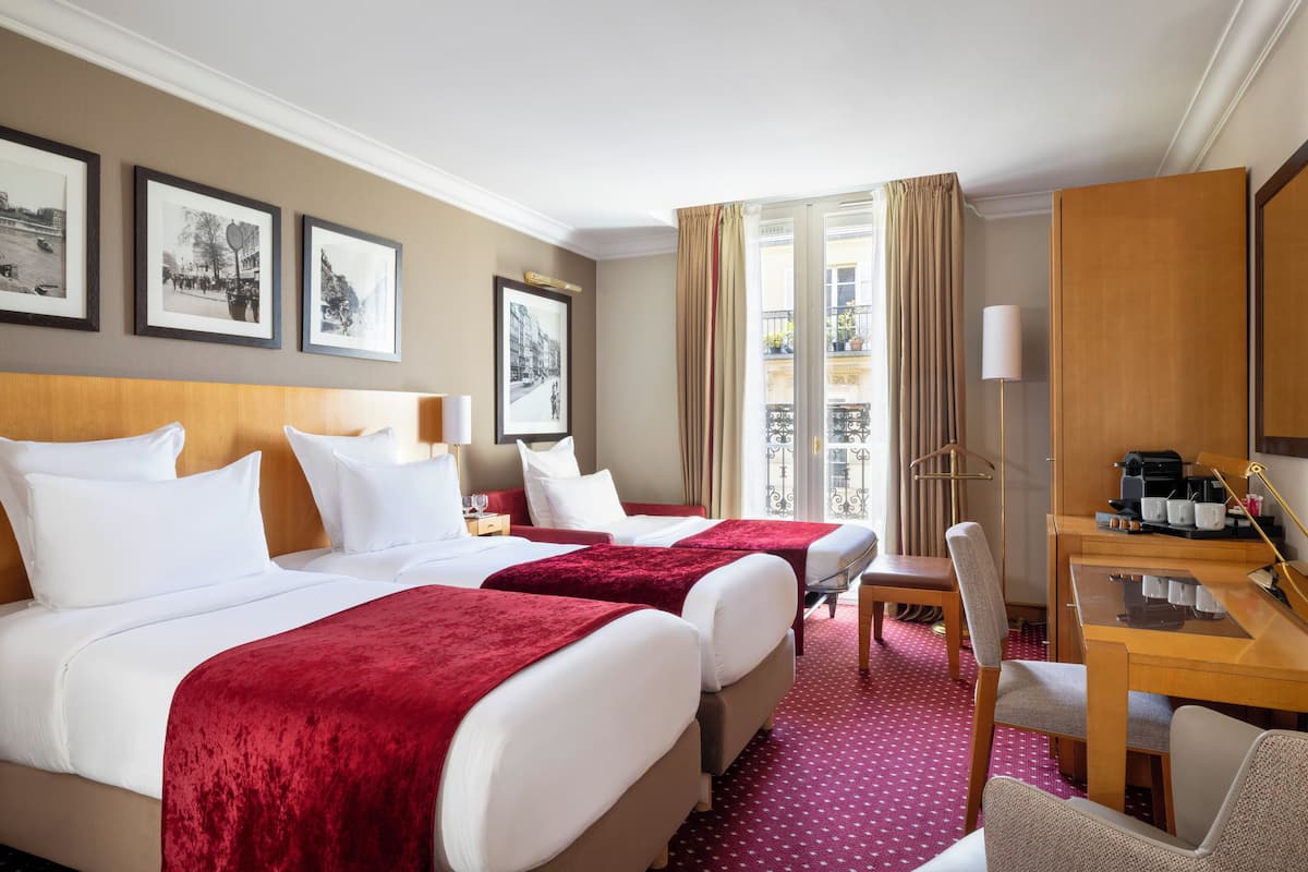 A hotel room with two beds, a desk, and a chair. There are framed pictures on the wall, a window with curtains, and a balcony view.