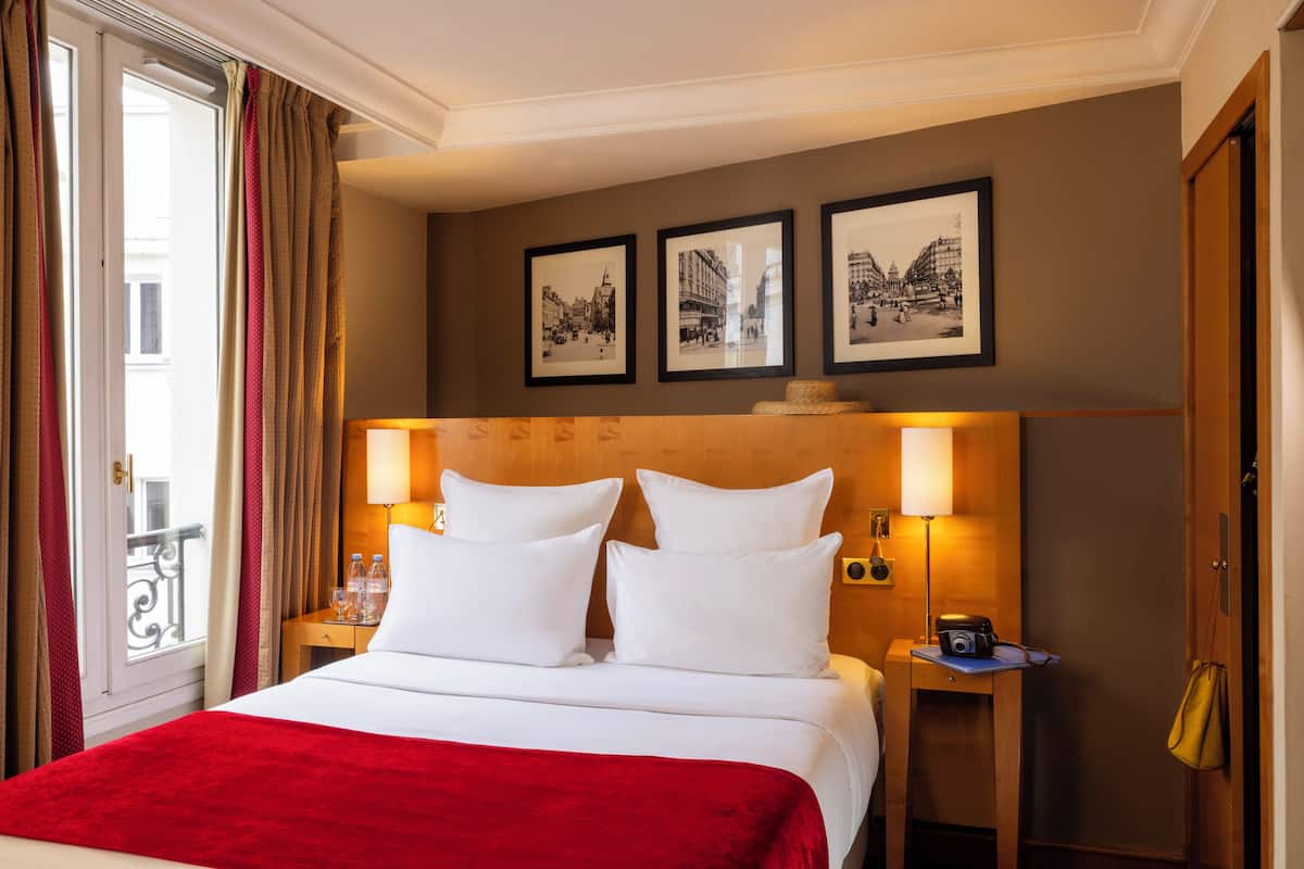 A hotel room with a bed, two bedside lamps, a wooden headboard, and three framed pictures on the wall.