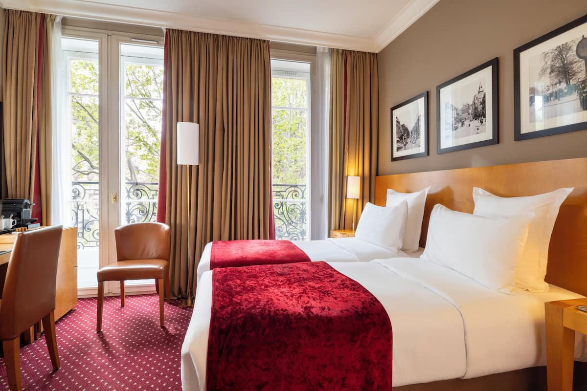 A hotel room with a bed, two chairs, a desk, and a balcony with curtains.