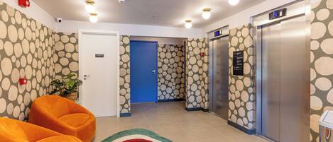 A hotel lobby with a reception desk, two orange armchairs, a blue door, and two elevators.