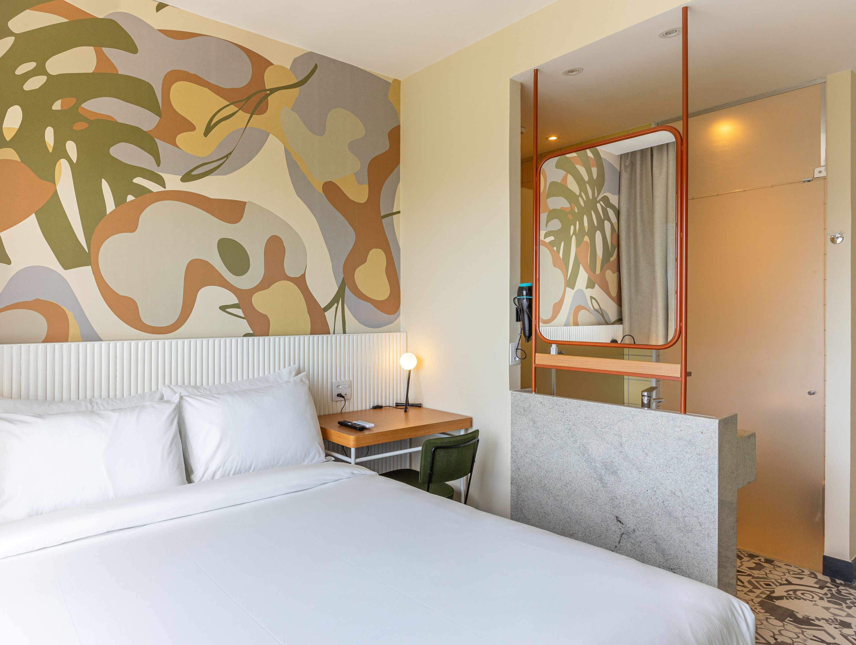 A hotel room with a bed, a desk, a chair, a mirror, and a bathroom visible through an open door.