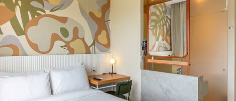 A hotel room with a bed, a desk, a chair, a mirror, and a bathroom visible through an open door.