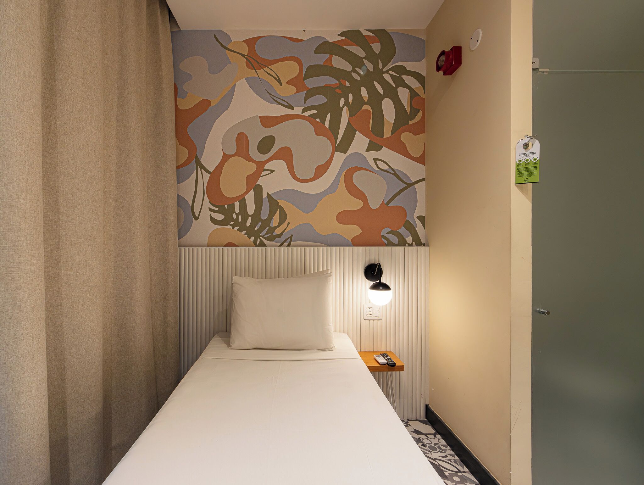 Una cama individual con colchón y almohada blancos, una lámpara fijada a la pared y una mesita de madera con un control remoto.