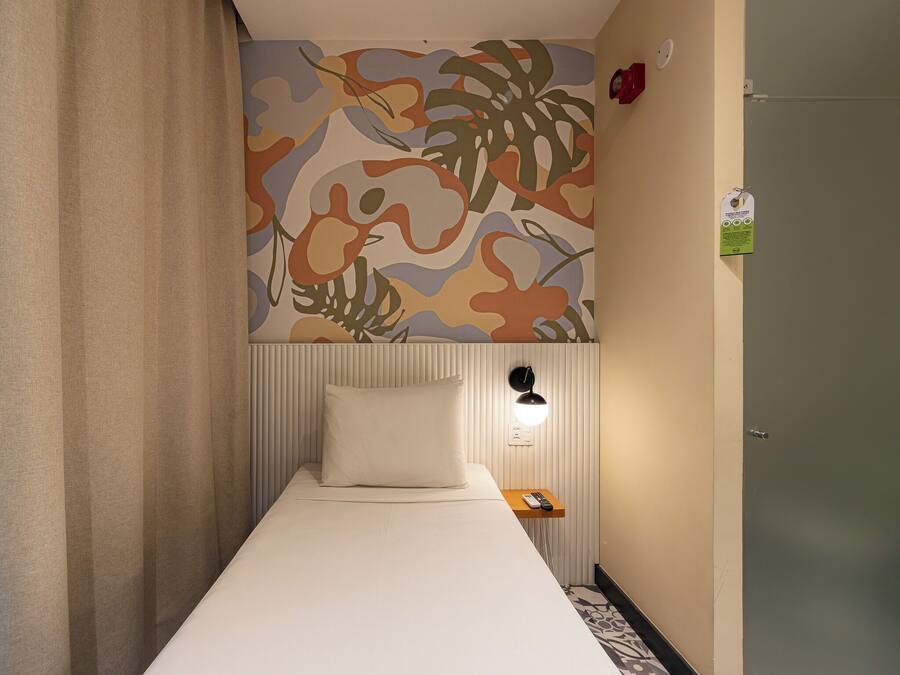 A single bed with a white mattress and pillow, a wall-mounted lamp, and a small wooden table with a remote control.