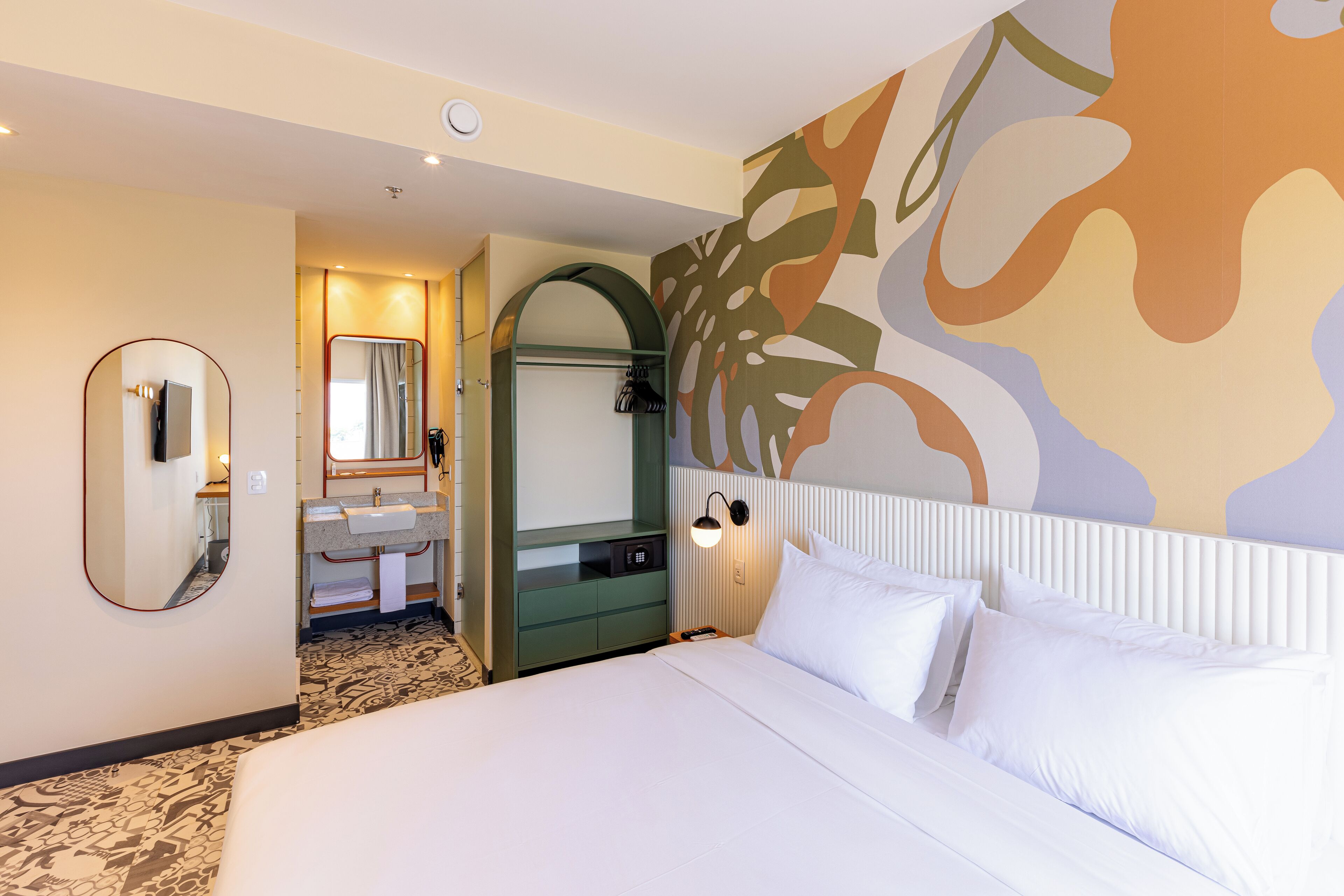 A hotel room with a large bed, a bathroom with a mirror and sink, and a wall with abstract art.