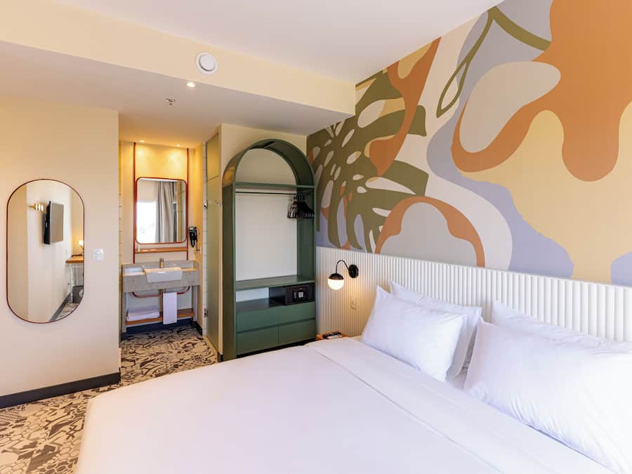 A hotel room with a large bed, a bathroom with a mirror and sink, and a wall with abstract art.