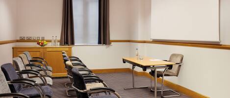 Une salle de conférence avec des rangées de chaises, un écran de projection, une table avec une bouteille et des verres, et une fenêtre avec des rideaux.