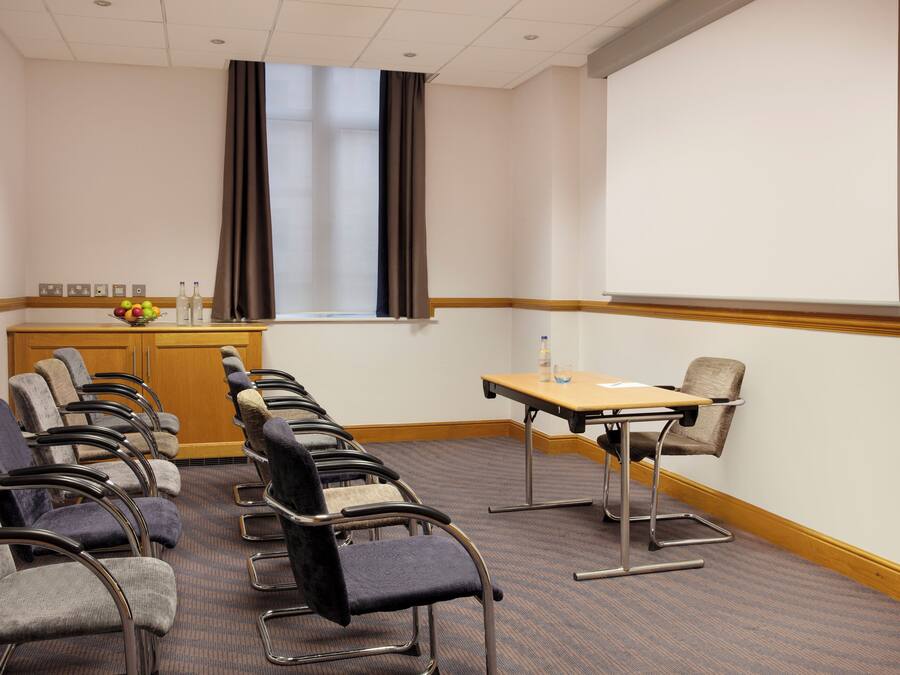 A conference room with rows of chairs, a projector screen, a table with a bottle and glasses, and a window with curtains.