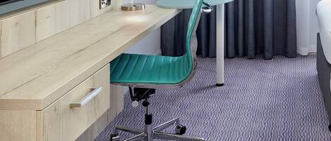 A hotel room with a wooden desk, a green office chair, a small round table, a bed, and a large window with curtains.