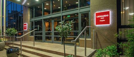 The exterior of the Leonardo Hotel in Croydon with a modern design, featuring a glass facade, a staircase leading to the entrance, and potted plants at the base.