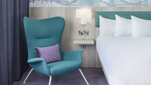 A teal armchair with a patterned pillow, a bedside table with a lamp, and a bed with white linens. - Leonardo Hotel London Croydon (Croydon)