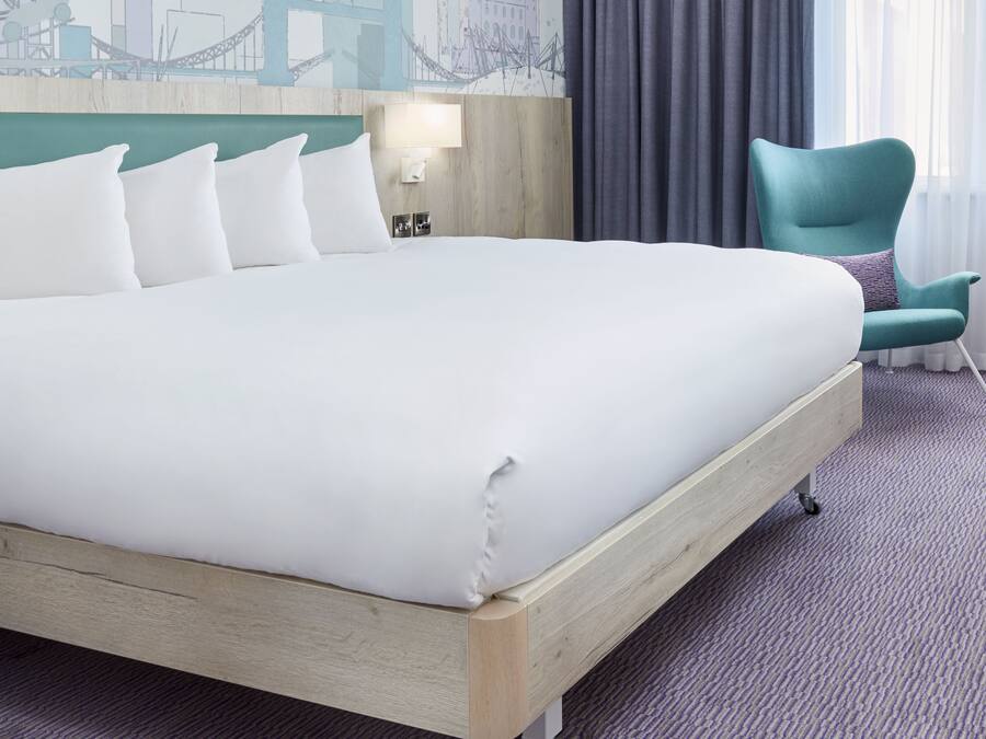 A hotel room with a large bed, a teal armchair, and a cityscape mural on the wall.