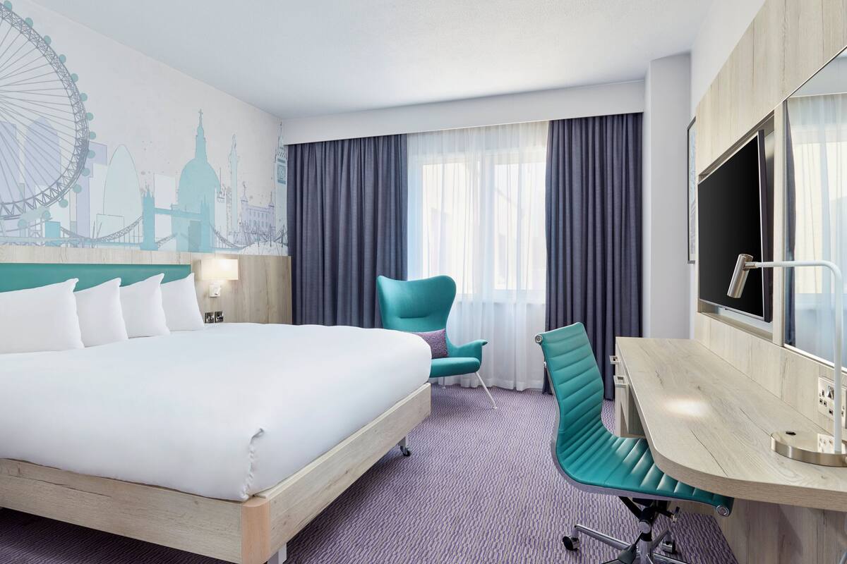 A hotel room with a large bed, a desk with a chair, a TV, and a mural of a city skyline.
