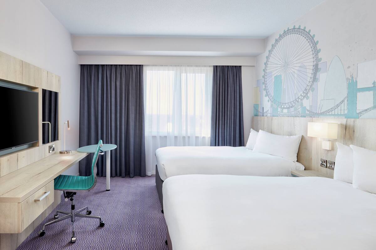 A hotel room with two beds, a desk with a chair, a TV, and a mural of a Ferris wheel.