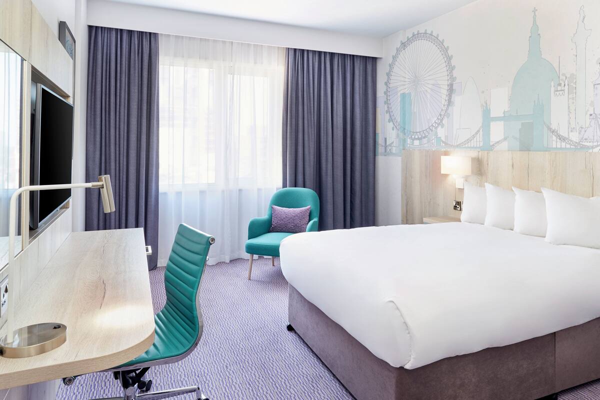 A hotel room with a bed, a desk, a chair, a large window with curtains, and a wall mural of a cityscape.