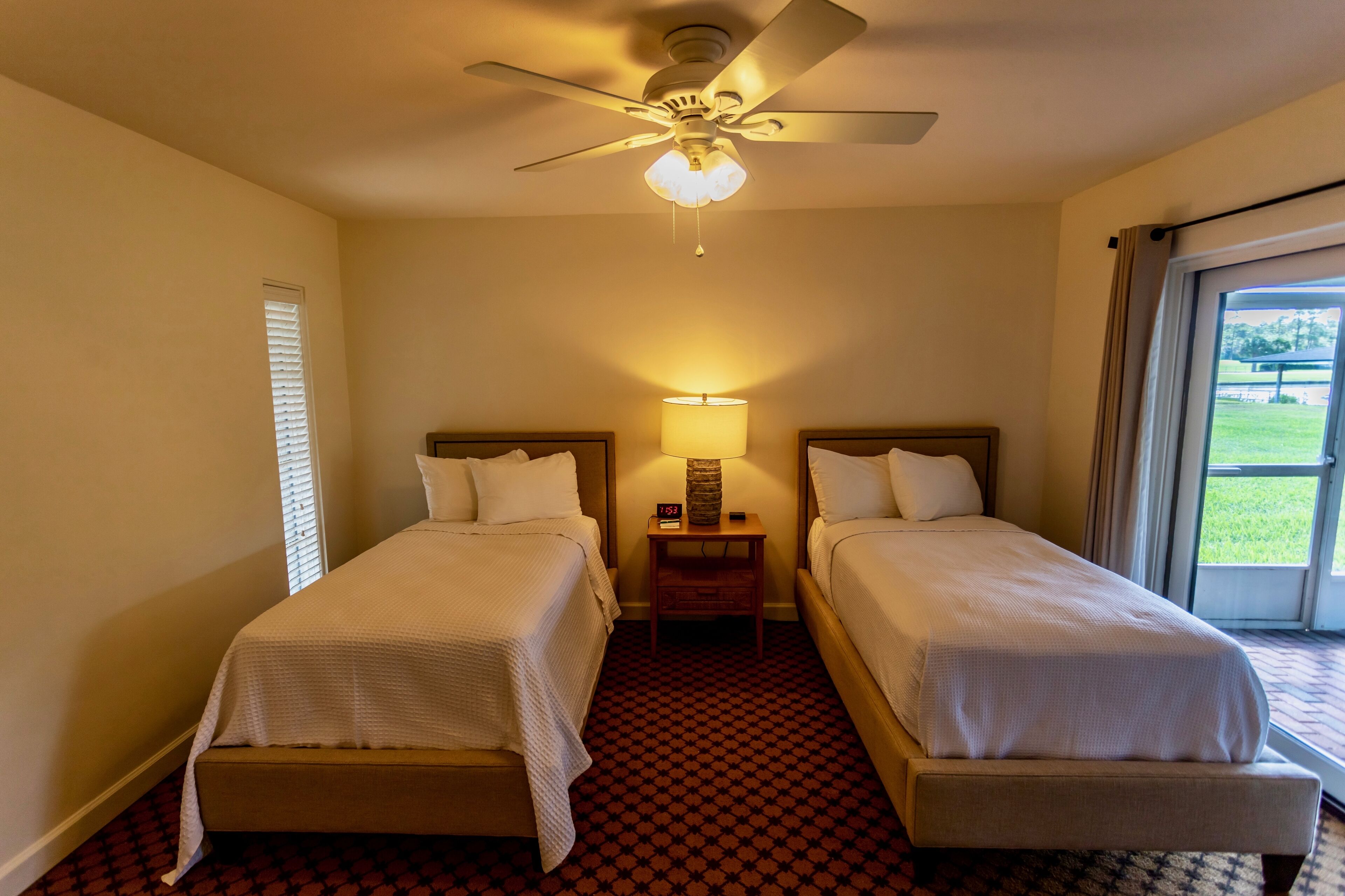 A hotel room with two single beds, a ceiling fan, a nightstand with a lamp, and a window with blinds.