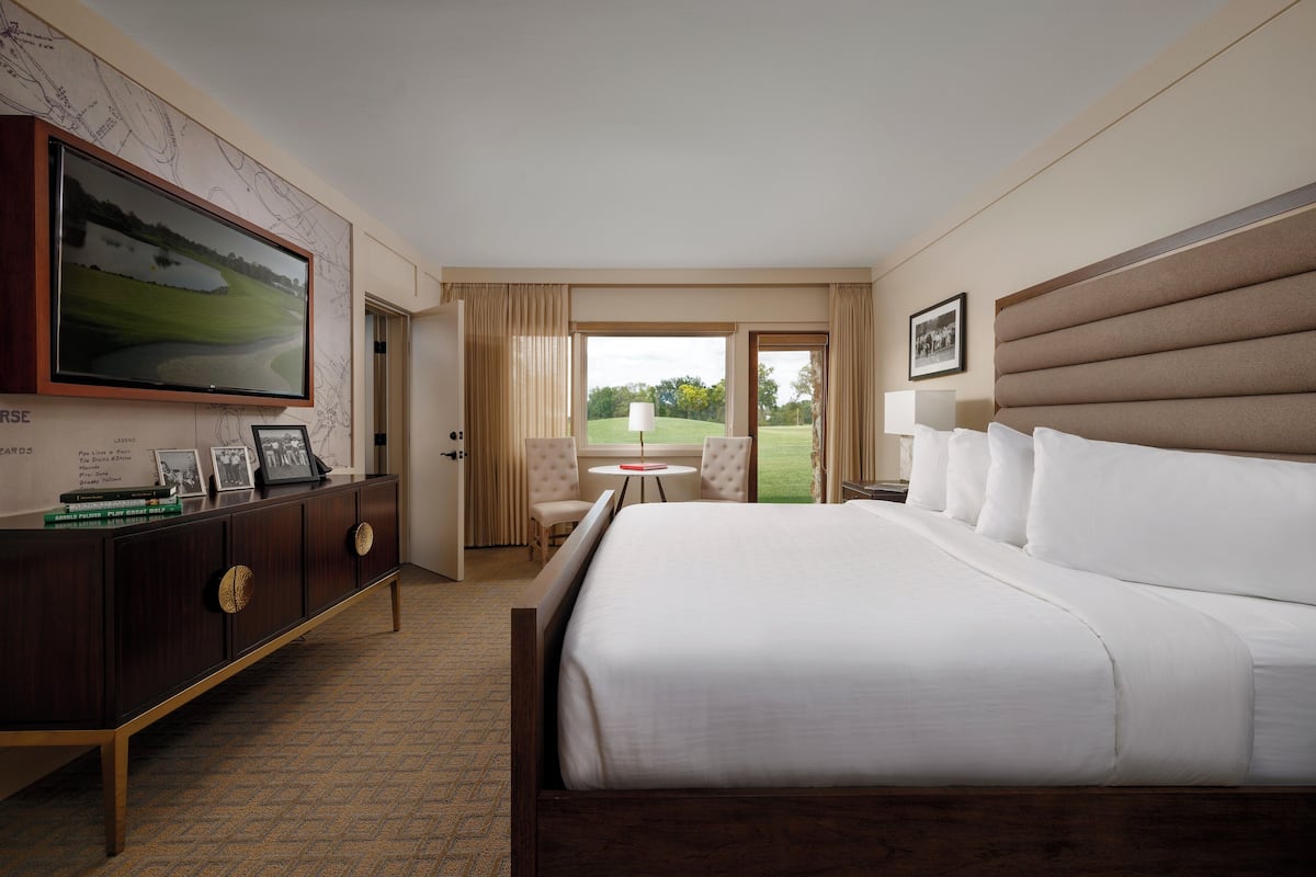 A hotel room with a large bed, a wooden dresser, a television mounted on the wall, and a view of a golf course through the window.