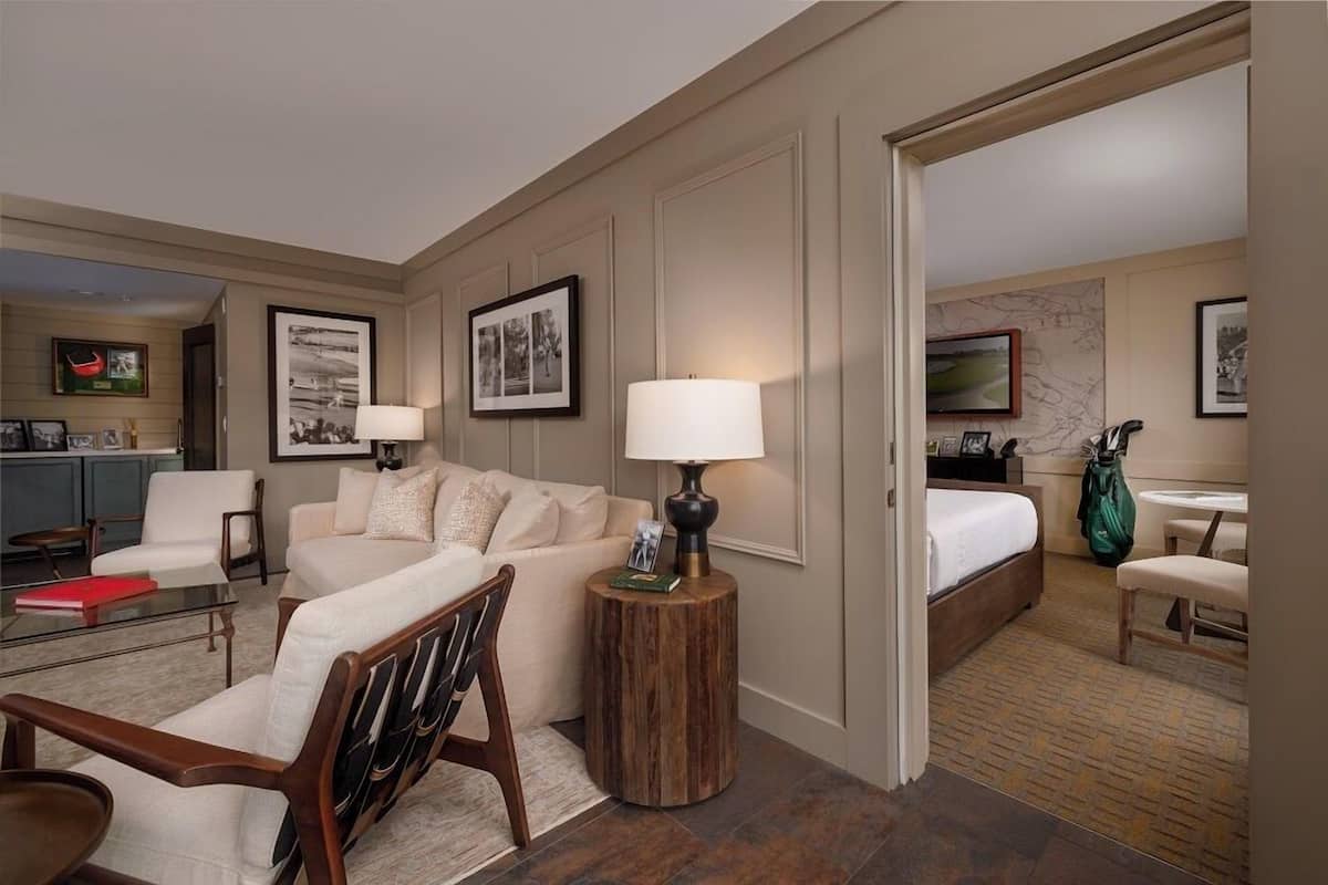 A hotel room with a living area, a bed, a small table with a lamp, and a view of the kitchen and bathroom through an open door.