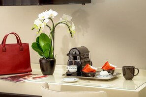 Ett hotellrum med ett glasbord, en röd handväska, en krukväxt, en kaffebryggare och ett bricka med koppar och en tekanna.