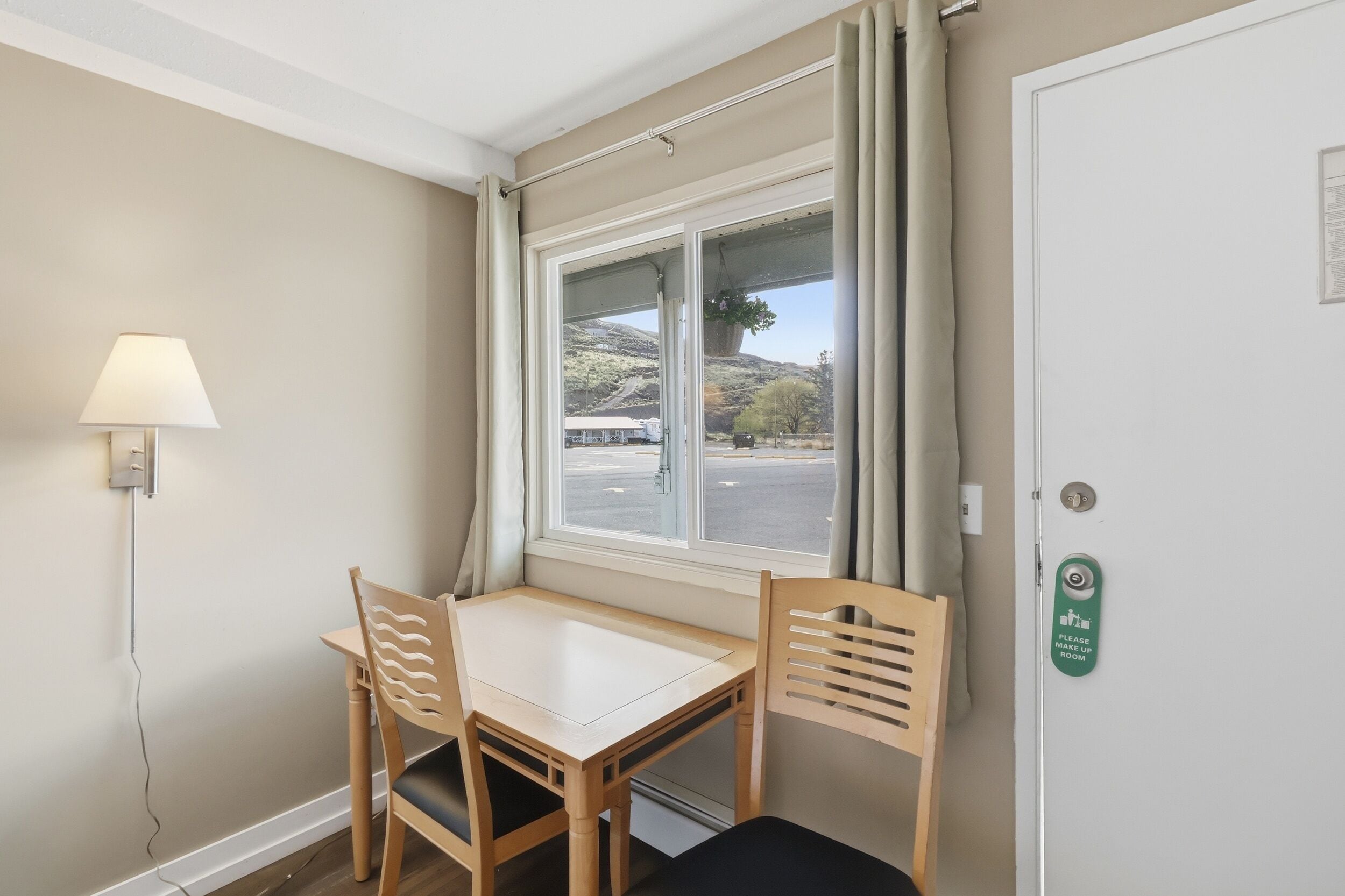 A room with a table and two chairs, a window with curtains, a lamp, and a door with a 'Please Make Up Room' sign.