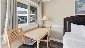 Ett hotellrum med en säng, ett bord, två stolar, ett fönster med gardiner, en lampa och en tavla på väggen.
