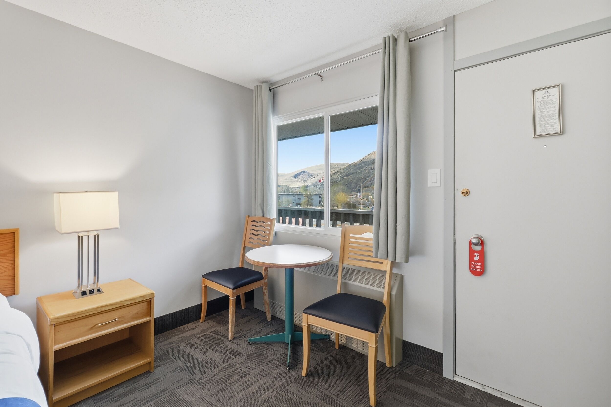 A hotel room with a bed, a bedside table with a lamp, a small round table with two chairs, a window with a view of mountains, and a door with a fire alarm.