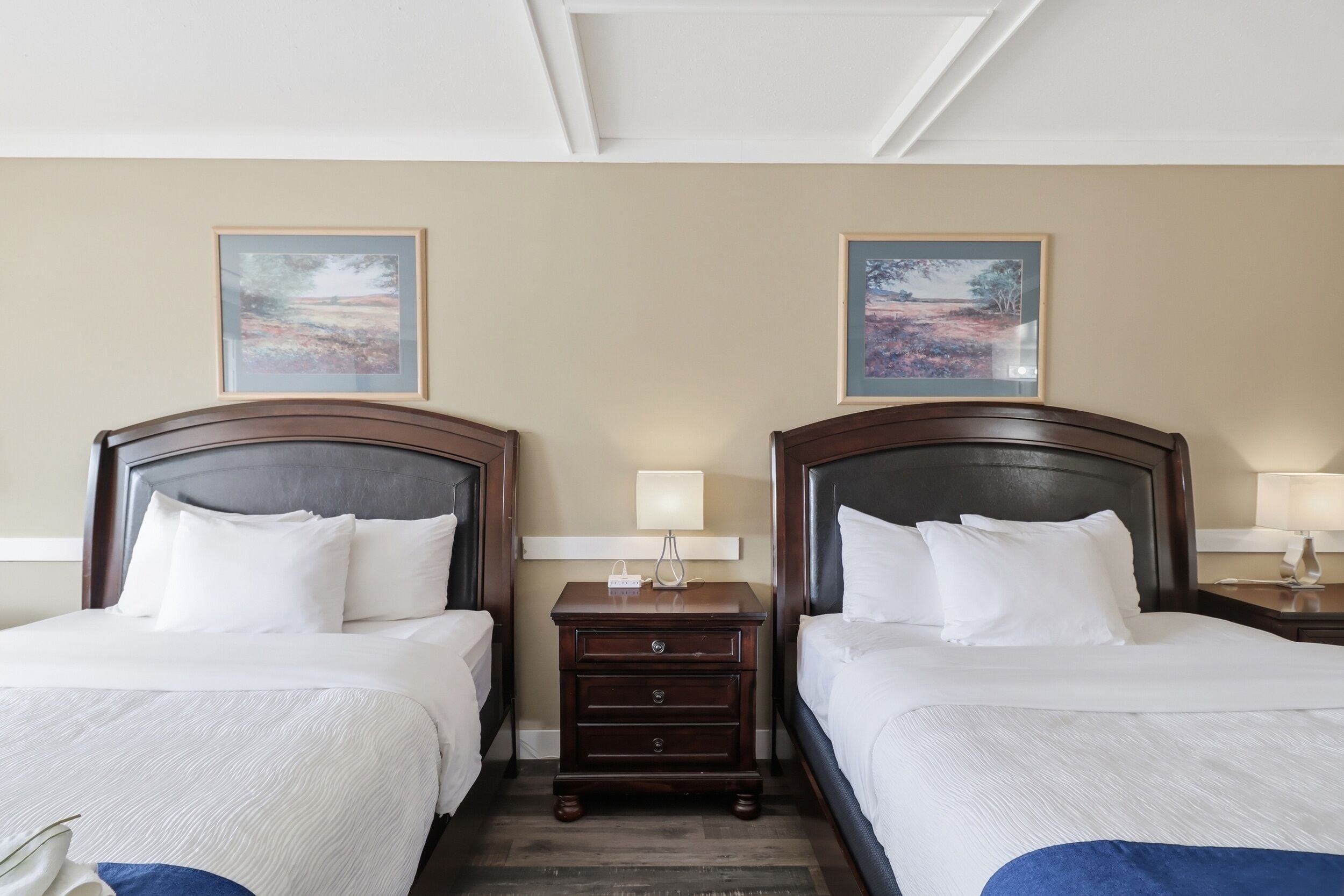 Une chambre d’hôtel avec deux lits, une table de chevet avec une lampe et deux tableaux encadrés au mur.