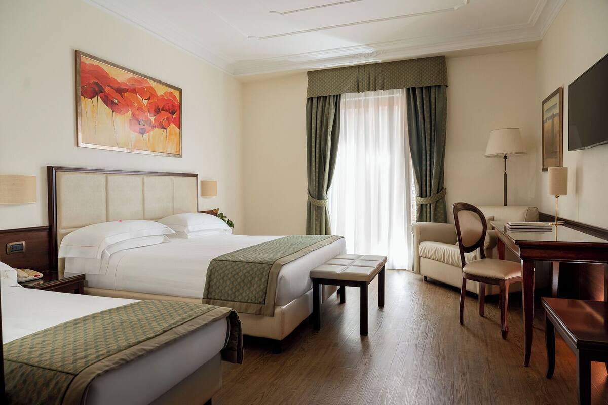 A hotel room with a bed, a desk, a chair, a bedside table, and a painting on the wall.