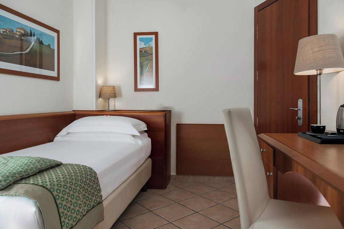 A hotel room with a bed, a desk, a chair, a lamp, and two framed pictures on the wall.