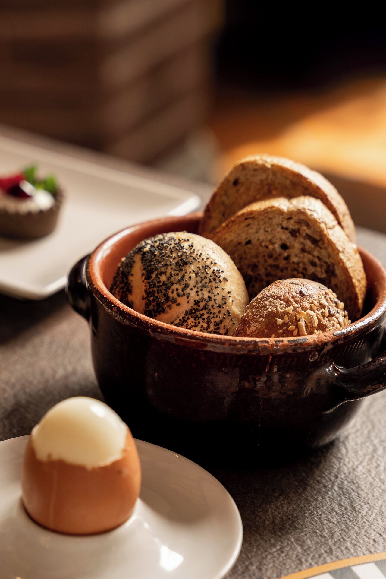 A bowl of bread with seeds, a small plate with a round item, and a blurred background.