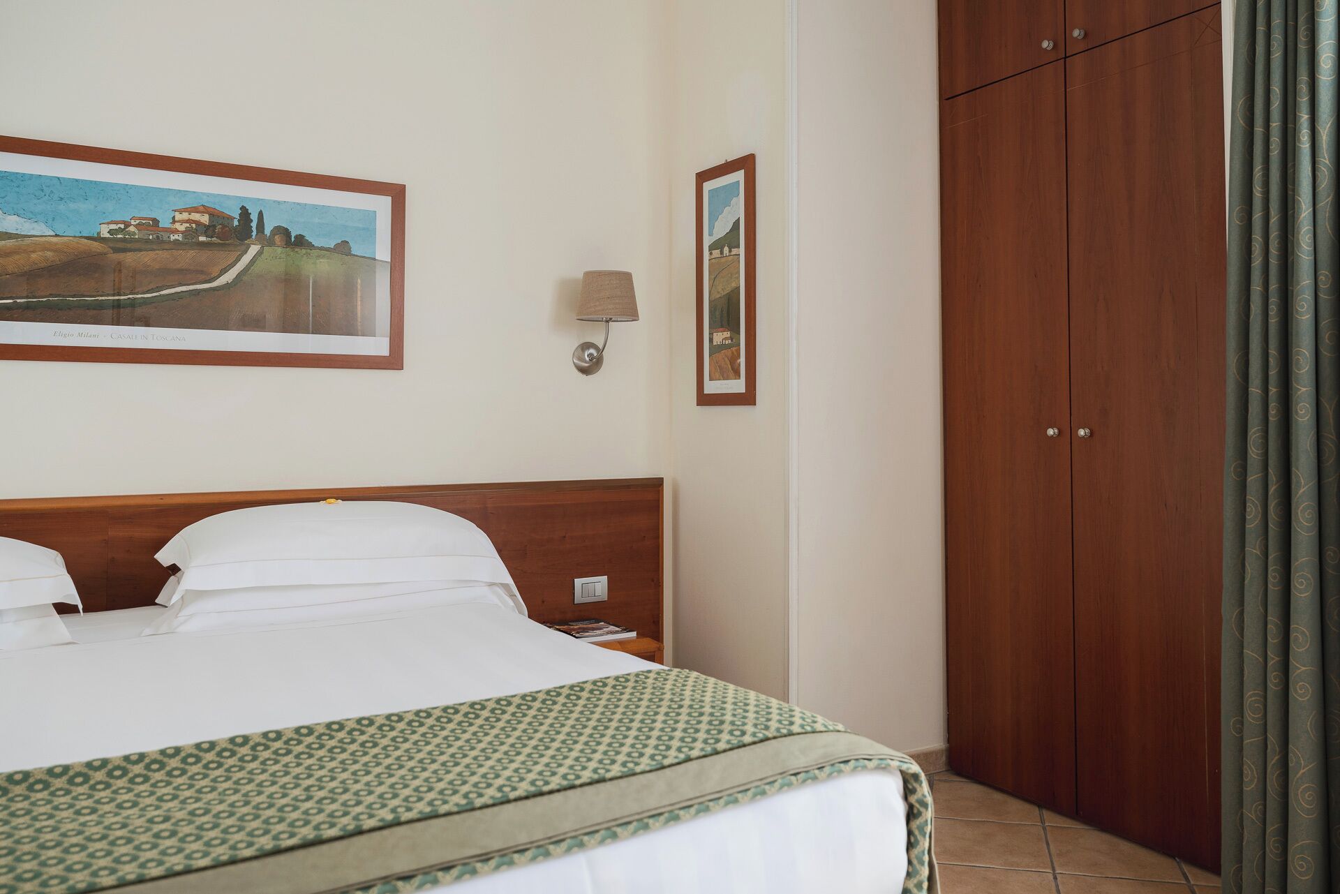 A hotel room with a bed, a nightstand, a lamp, a wardrobe, and two framed pictures on the wall.